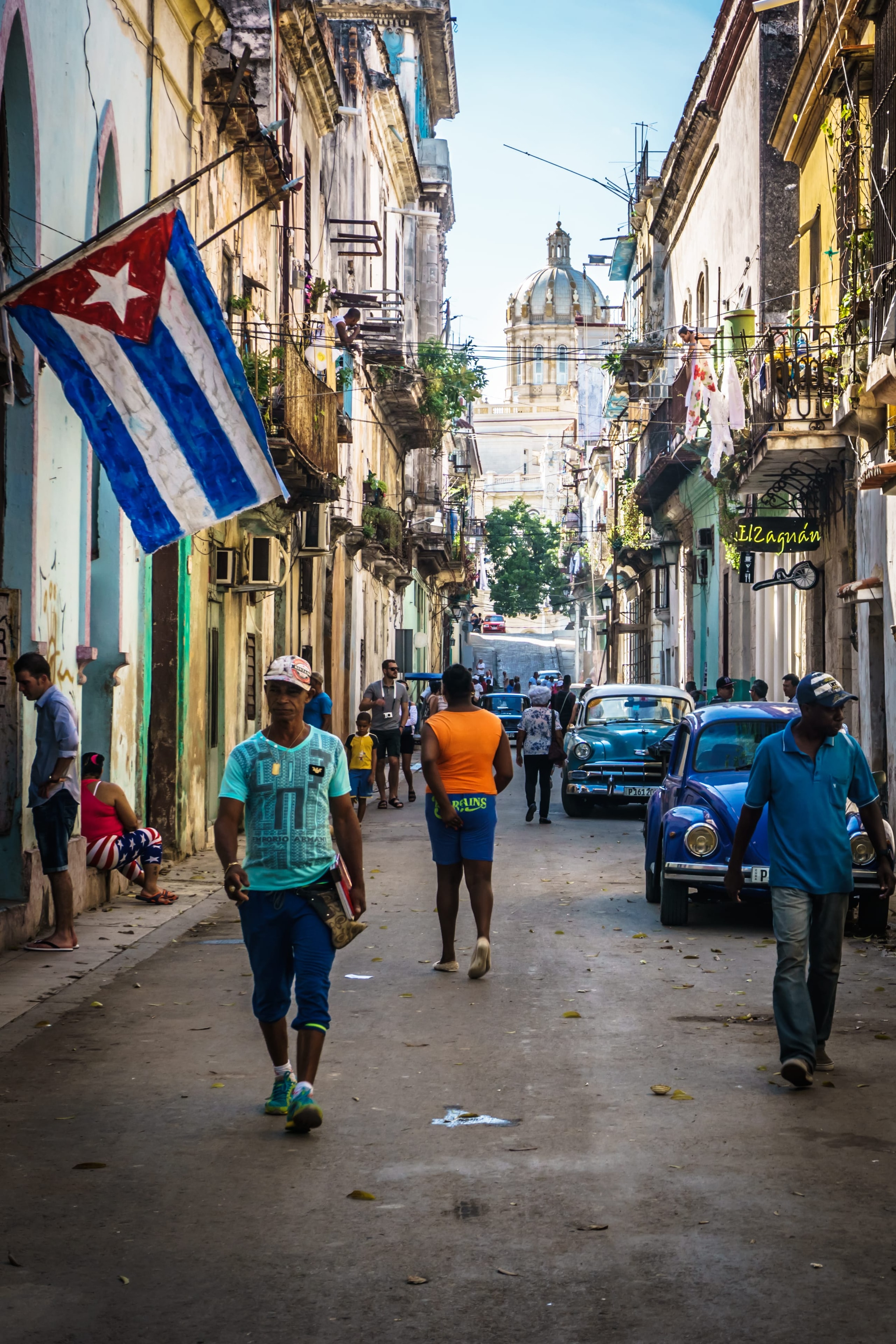 Busy Havana street with Cuban flag hanging from balcony, pedestrians, blue vintage cars, and golden cathedral dome in background, Cuba