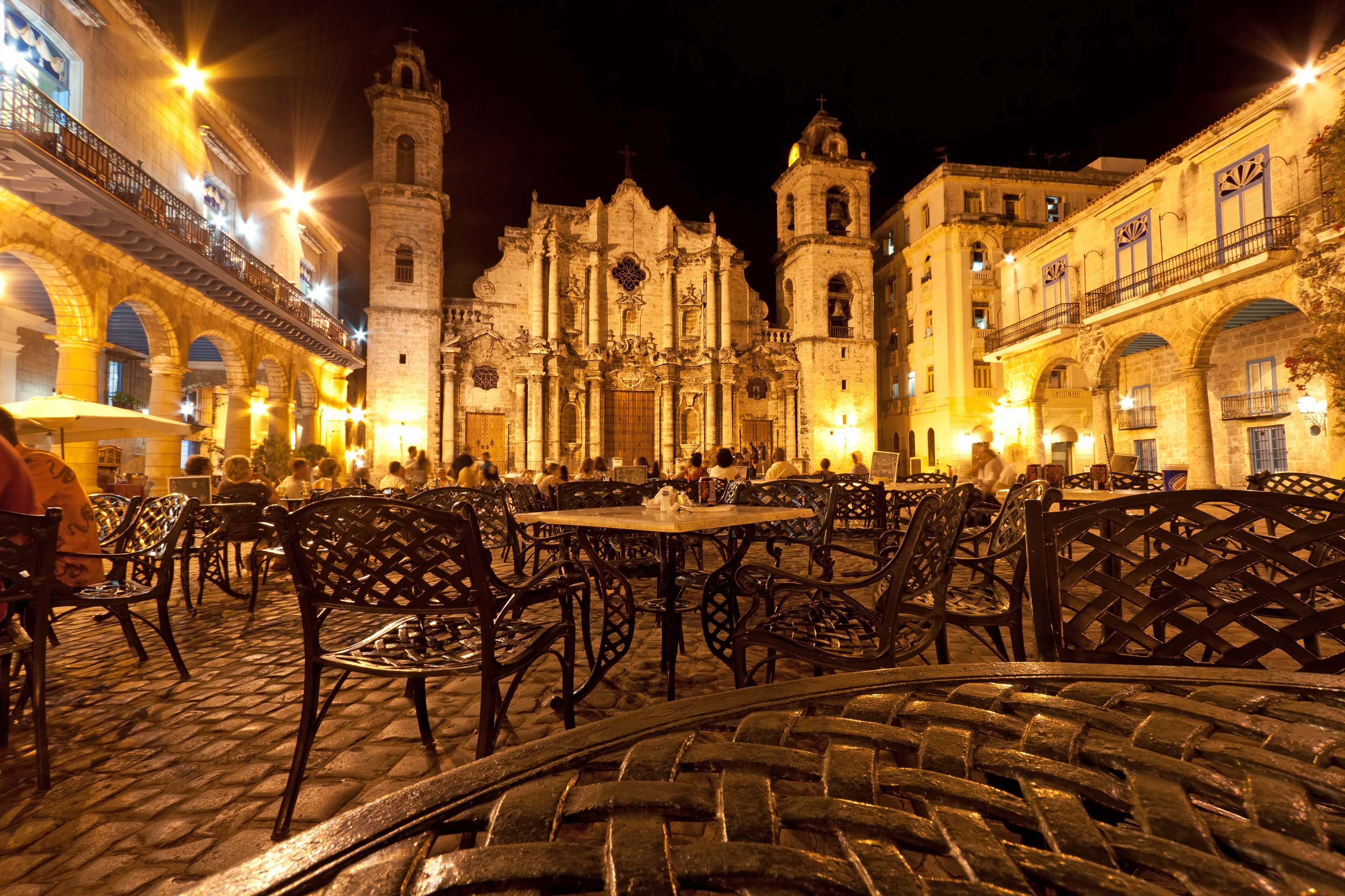 Plaza de la Catedral Havana at night with baroque cathedral illuminated in gold, iron café tables on cobblestones, and diners, Cuba