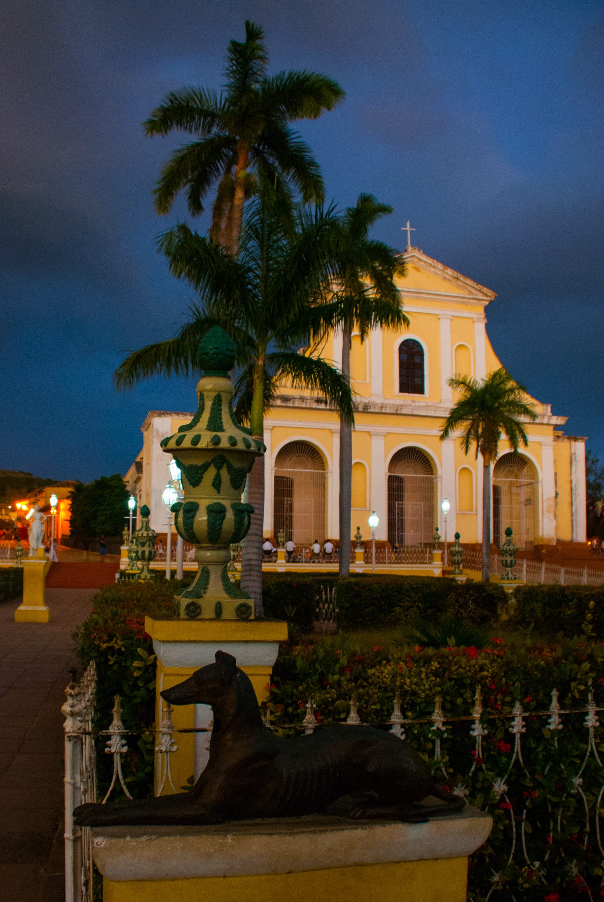 Plaza Mayor Trinidad at night with yellow Iglesia de la Santísima Trinidad illuminated, iron fence, bronze dog sculpture, and royal palms under a stormy sky, Cuba
