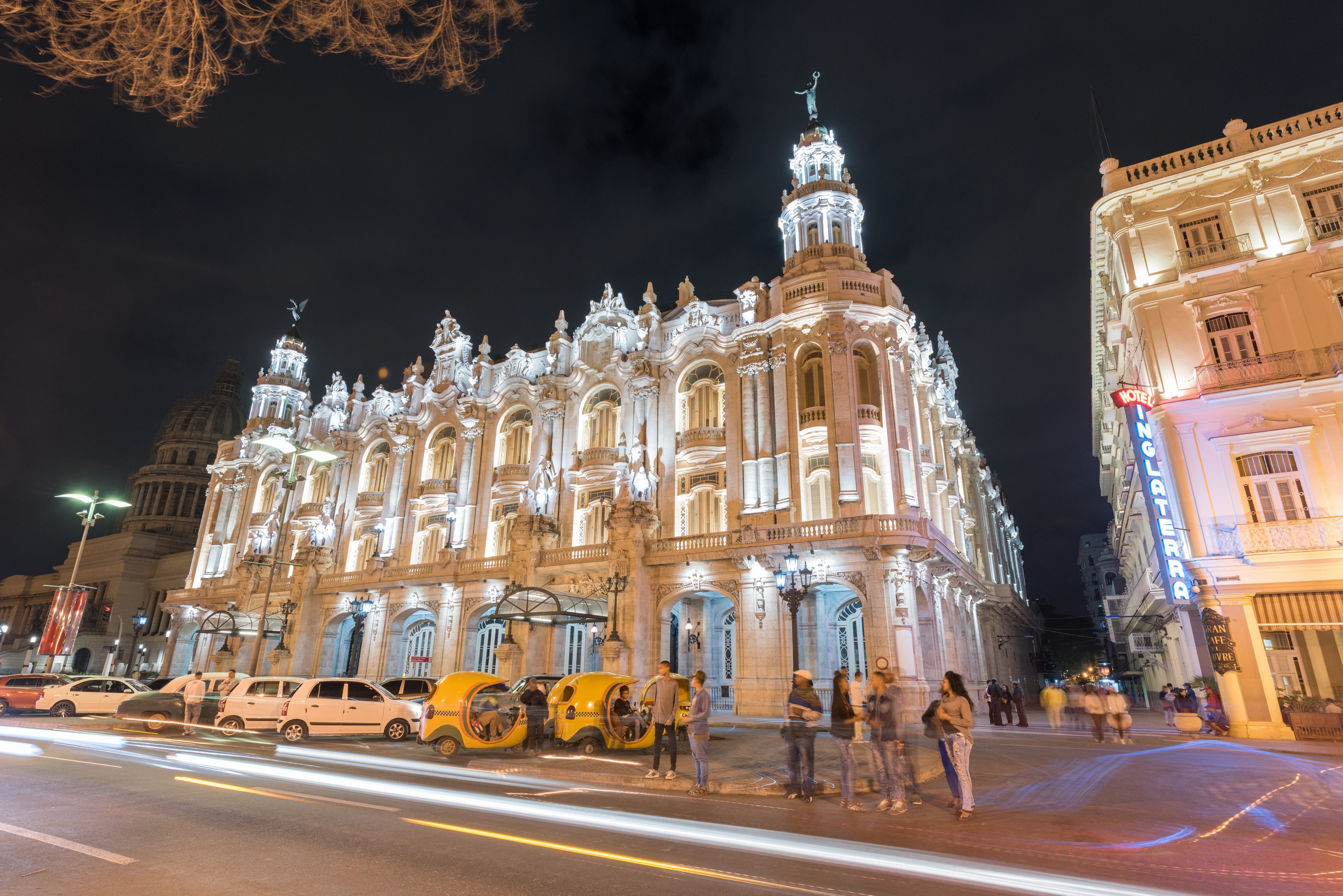 Gran Teatro de La Habana illuminated at night with baroque white facade, angel statues, yellow coco-taxis, pedestrians, and light trails on the wet street, Cuba