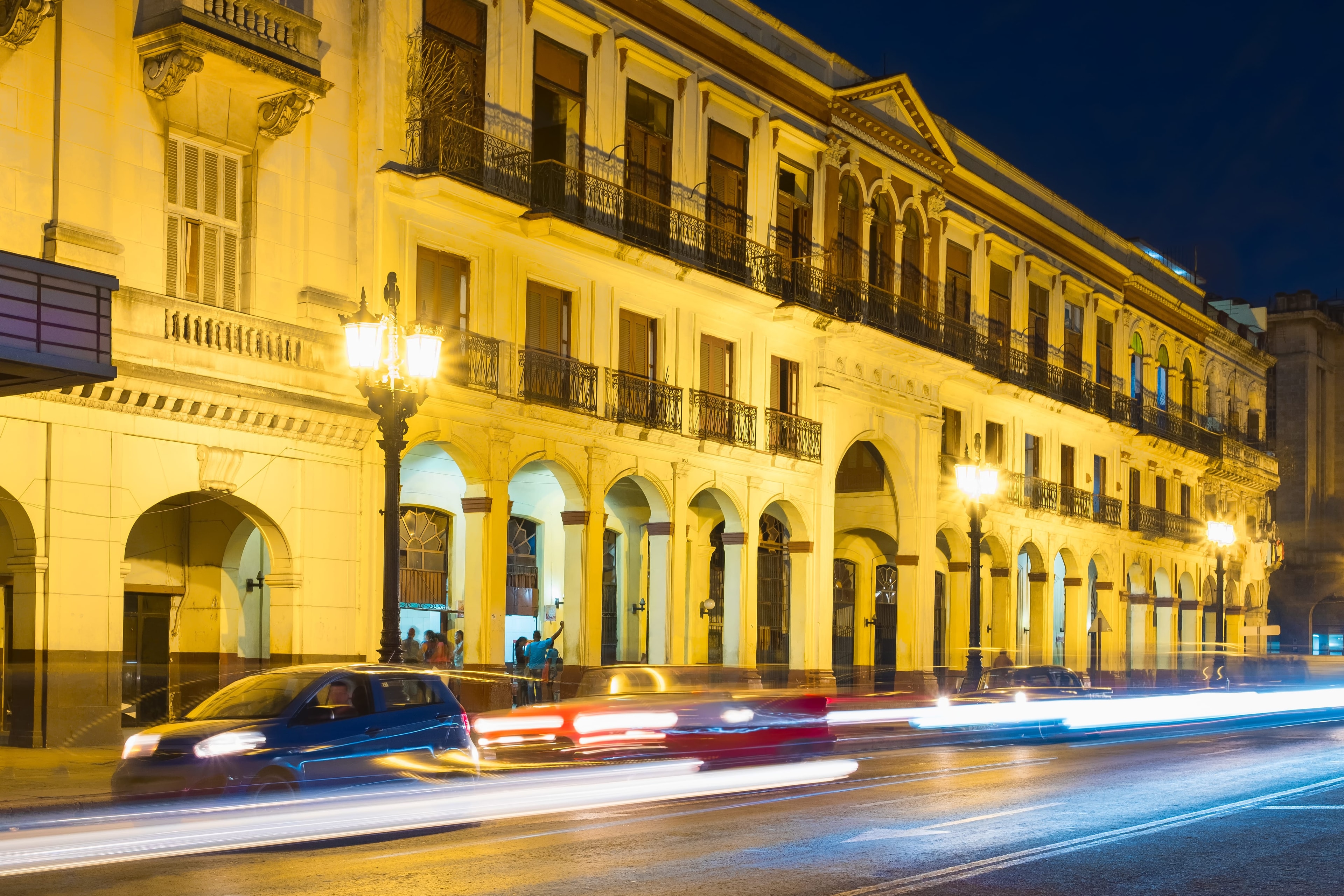 Old Havana neoclassical arcade at night with long-exposure light trails from passing cars and golden street lamps illuminating the colonnade, Cuba