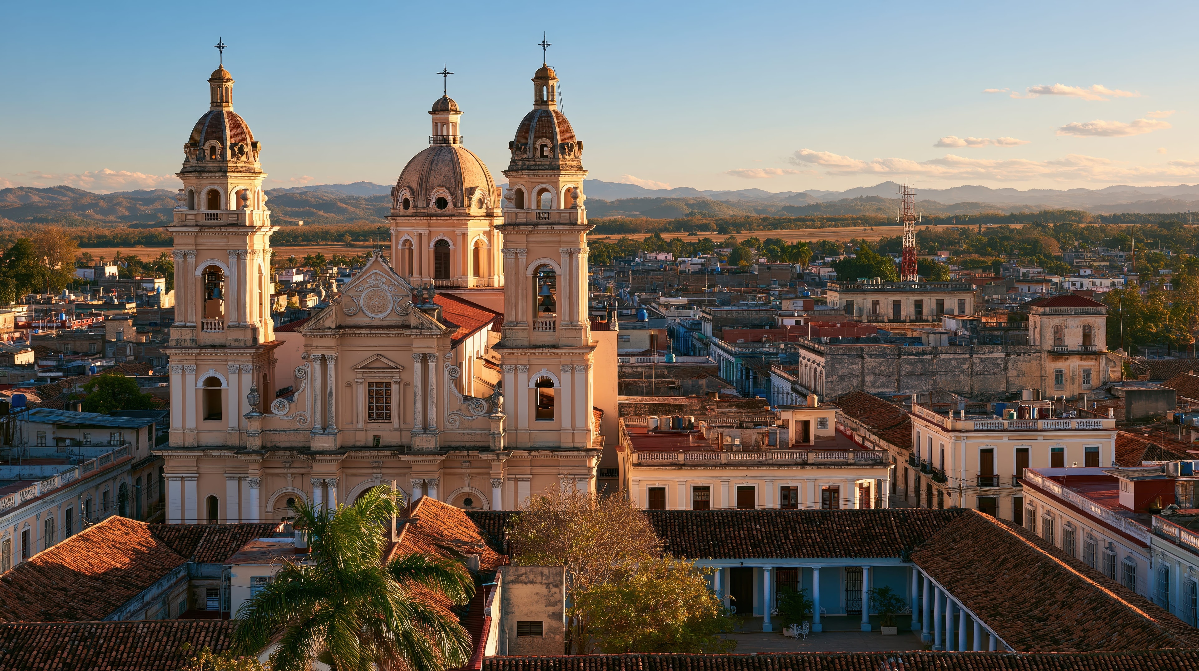Aerial golden hour view of baroque cathedral with twin towers rising above Camagüey rooftops and mountains, Cuba