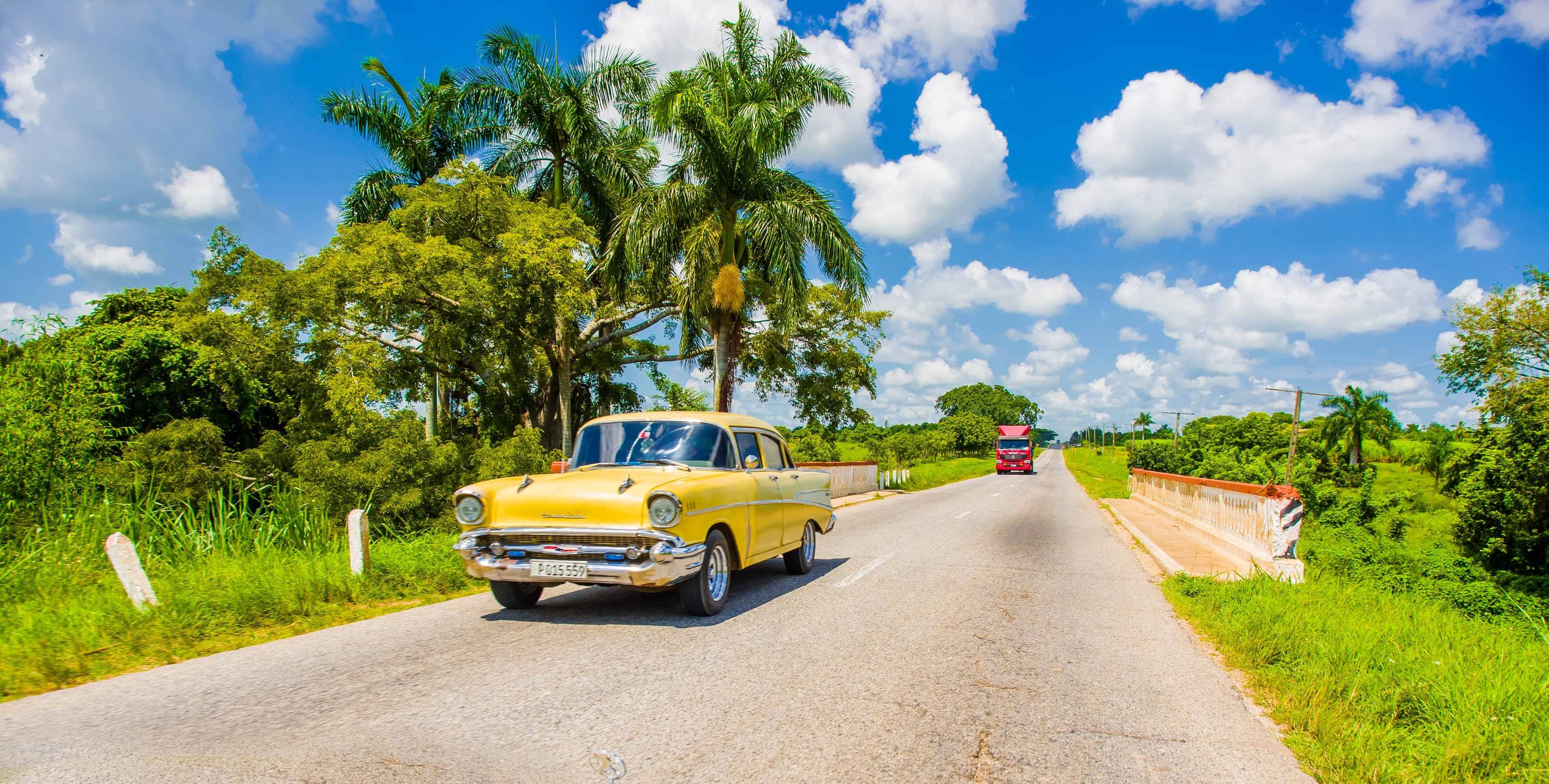 Yellow vintage Chevrolet driving on open Cuban highway through lush tropical landscape